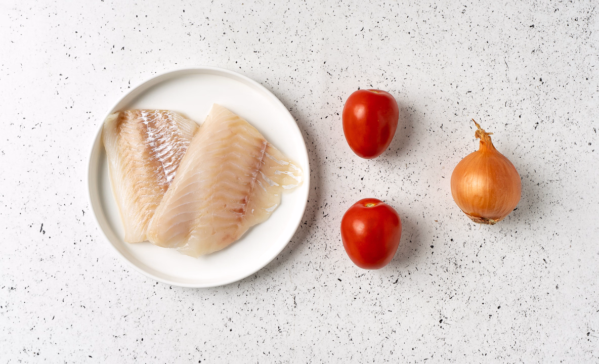 На фото два филе трески лежат на тарелке, справа от них — два небольших помидора, еще правее — неочищенная луковица. Вид сверху.