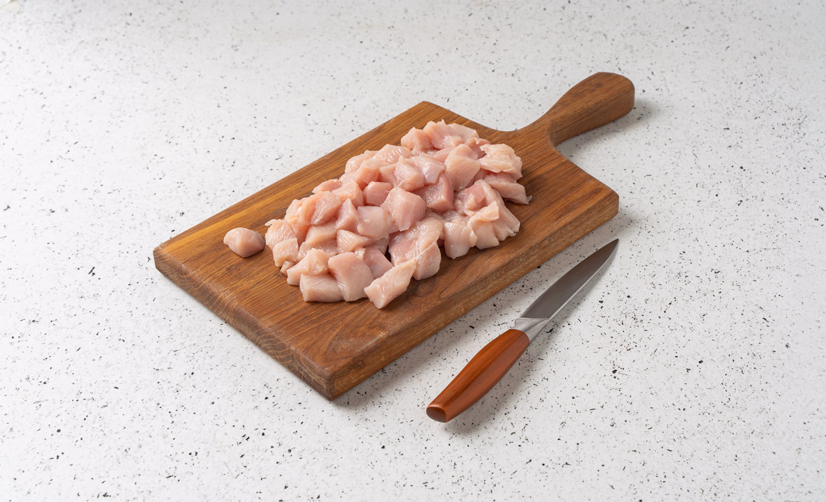 Сырые кусочки курицы нарезаны кубиками, лежат на резделочной доске, рядом нож