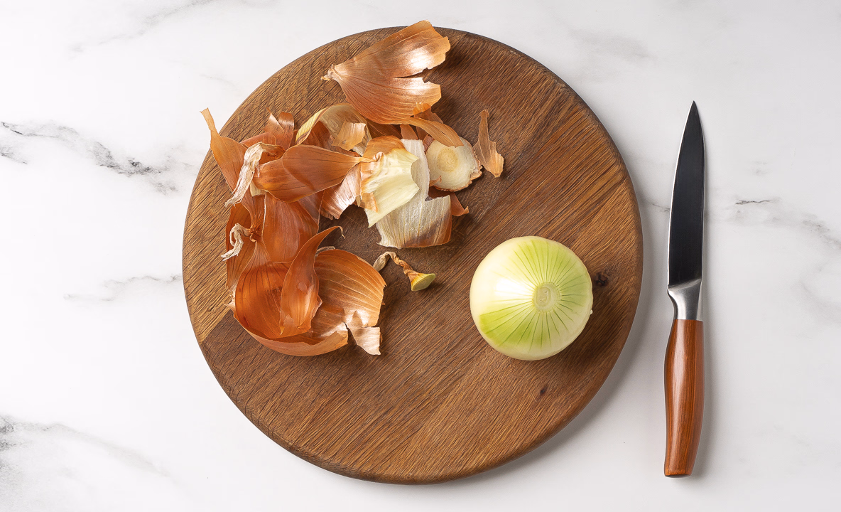 Очищенный лук лежит на деревянной доске. Рядом с луковицей лежит шелуха.