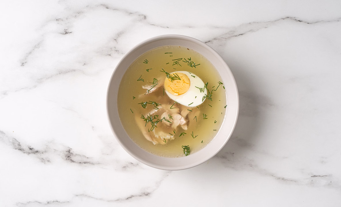 На столе стоит тарелка с бульоном, яйцом и куриным мясом. Бульон аккуратно посыпан небольшим количеством зелени.